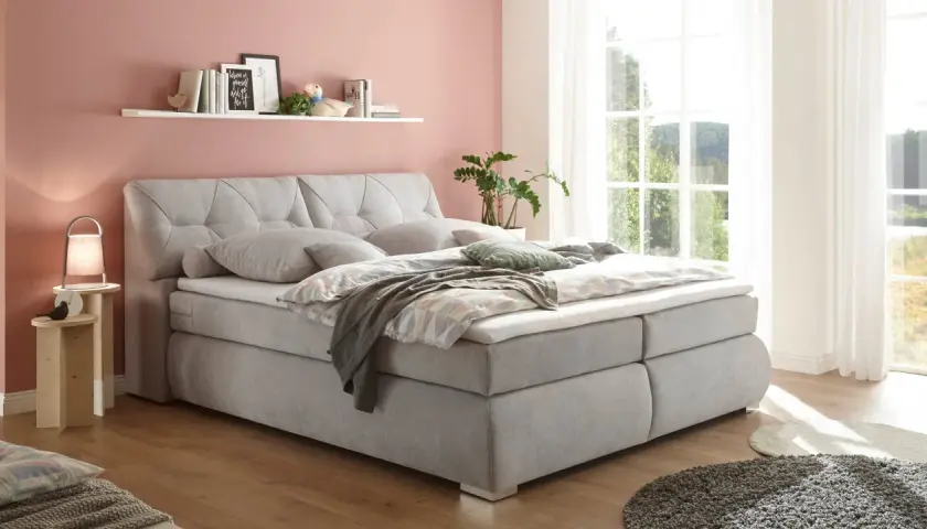 Ein hellgraues Boxspringbett mit Kissen und Decken lädt zum Entspannen ein. Die Wand in Rosé und das Fenster mit Blick ins Grüne schaffen eine harmonische Atmosphäre für ein **Schlafzimmer Feng Shui**.