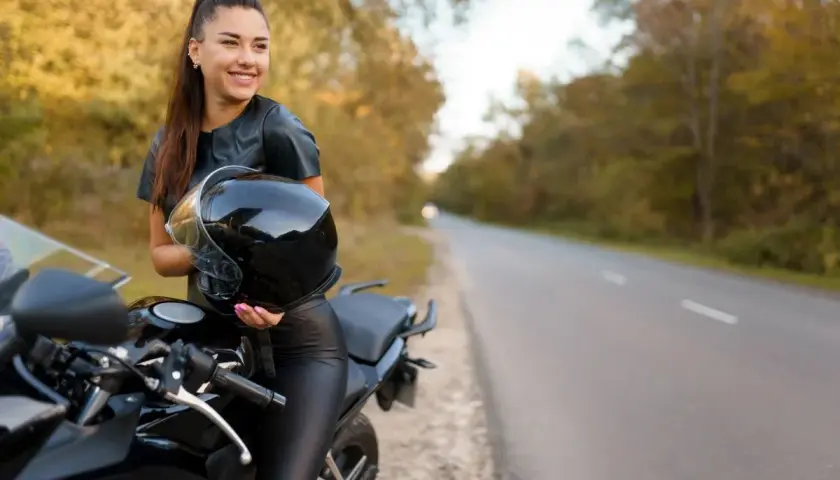 Uśmiechnięta kobieta w skórzanym stroju trzyma kask obok czarnego motocykla. Idealne motory dla kobiet na jesienną przejażdżkę.