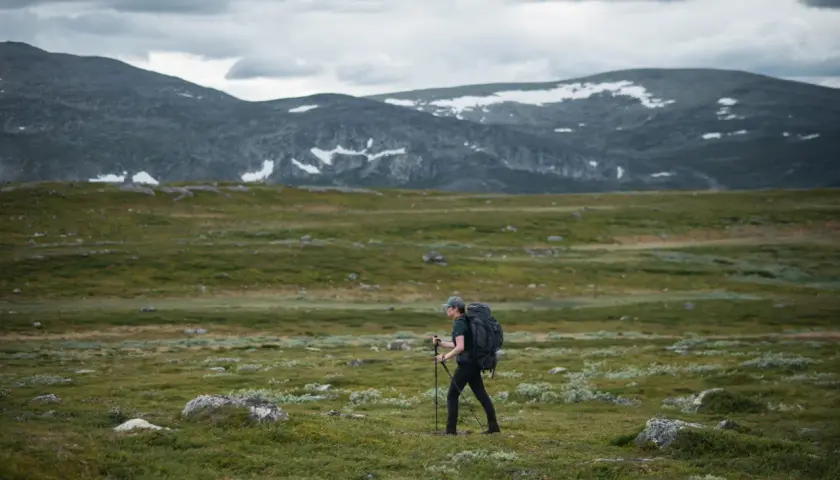 Vandrare med ryggsäck och stavar går över kalfjäll med snöfläckar på berg i bakgrunden. En helags karta skulle visa vägen.