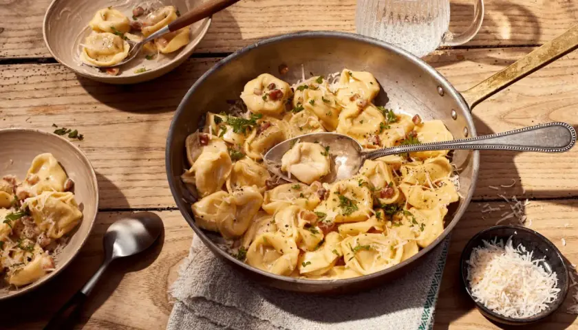 Herzhafte Tortellini Carbonara, garniert mit Petersilie und Parmesan, serviert in einer Pfanne und Schalen auf Holztisch.