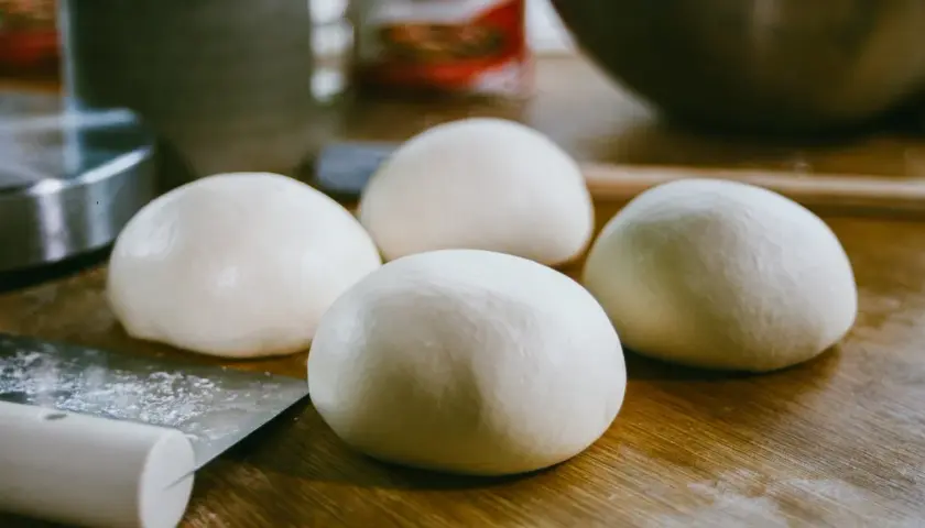 Cztery kulki surowego ciasta na pizzę leżą na drewnianym blacie obok noża.
