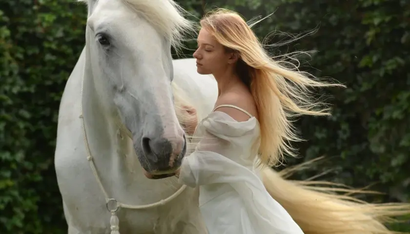 Una joven rubia con un vestido blanco se inclina hacia un majestuoso caballo blanco. Es como soñar con caballos, una escena etérea.