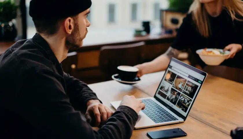 Mężczyzna w czapce pracuje na laptopie, analizując dane. Obok stoi filiżanka kawy. To praca ux researchera.