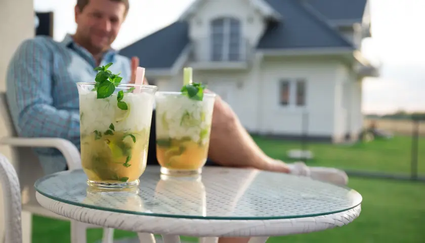 Jak przygotować drinki w dzbankach na domową imprezę? Dwa orzeźwiające mohito z miętą i limonką na stoliku, w tle mężczyzna i dom.