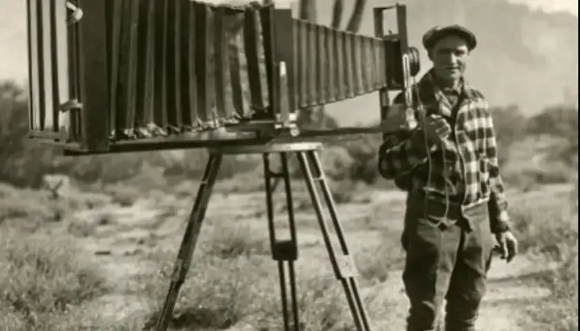 Mężczyzna w czapce i kratowanej koszuli pozuje obok ogromnego, zabytkowego aparatu fotograficznego na statywie. To obraz z czasów pierwszych aparatów fotograficznych.