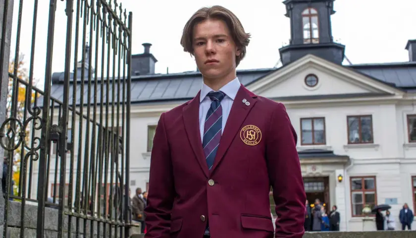 En ung man i skoluniform, som påminner om unga kungligheter, står framför en ståtlig byggnad.