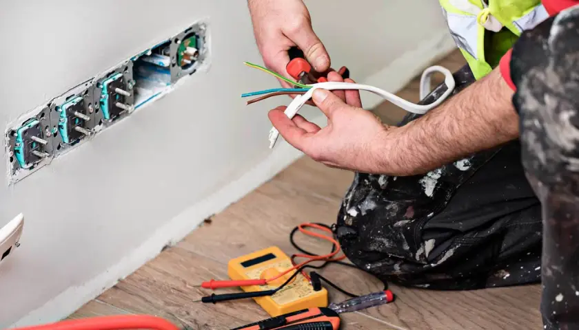 Elektryk przygotowuje kabel czarny, brązowy, niebieski do podłączenia gniazdek. Narzędzia i miernik leżą na podłodze.