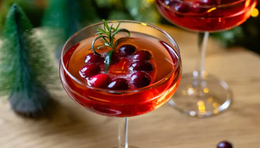 Festliche Aperol-Cocktails mit Cranberries und Rosmarin, perfekt für Aperol Weihnachten. Kleine Bäume und Lichter im Hintergrund.