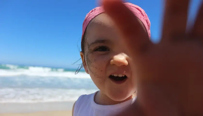 Niña con bandana rosa y arena en la cara, disfrutando de sus viajes con niños en la playa.