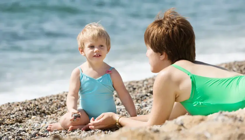 Mama i dziecko cieszą się wakacjami nad morzem z dziećmi. Dziewczynka w niebieskim kostiumie bawi się kamyczkiem, a mama w zielonym leży obok.