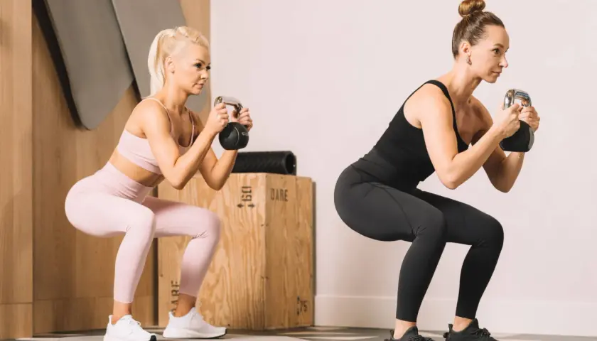 Dwie kobiety wykonujące przysiady z kettlebellami w siłowni.