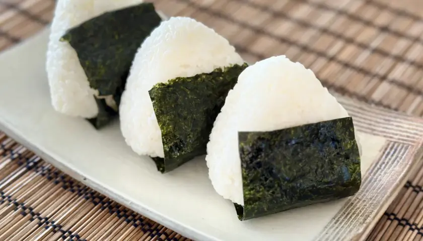 Three triangular onigiri, made with fluffy sushi rice and wrapped in dark green nori seaweed, sit on a white plate.