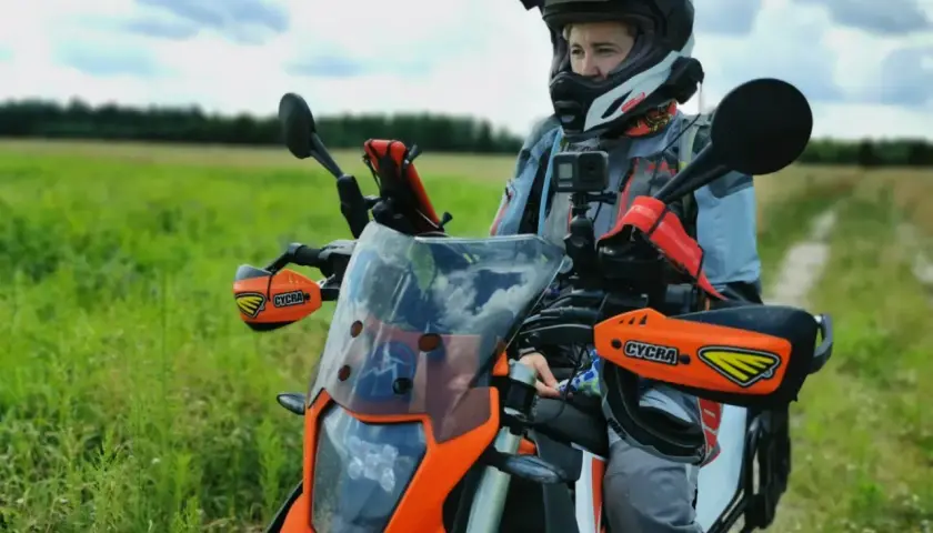 Motocyklista w kasku siedzi na pomarańczowym motocyklu w terenie, na tle zielonego pola i nieba.