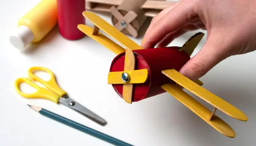 Un avión hecho con materiales de **avión manualidades**, sostenido por una mano. Tijeras y lápiz cerca, listos para crear más.