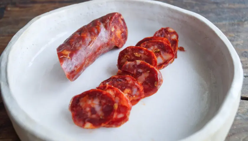 Ein Stück und Scheiben einer würzigen Salsiccia Wurst liegen in einer weißen Schale auf einem Holztisch.