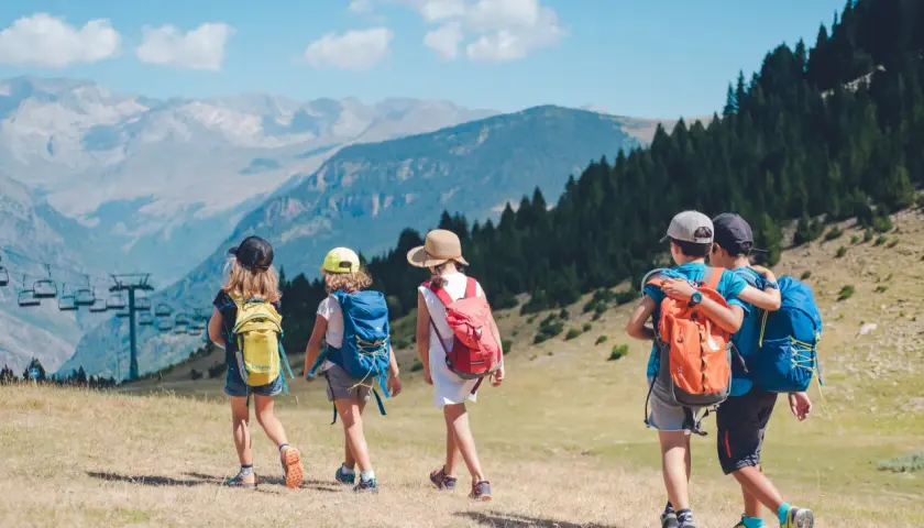 Niños explorando los Pirineos con mochilas, disfrutando de un día soleado con vistas a las montañas y un telesilla.