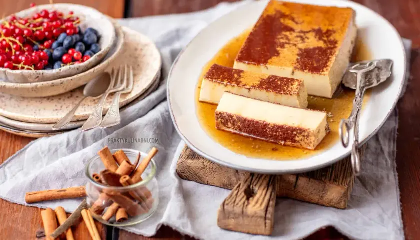 Pyszny creme caramel na talerzu, polany syropem. Obok miseczka z owocami i laska cynamonu.