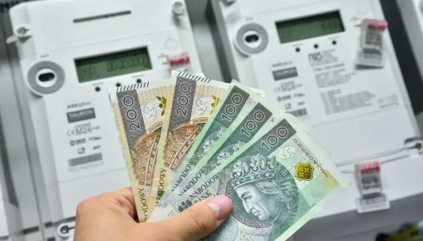 Dłoń trzyma polskie banknoty przed licznikami energii elektrycznej. Zastanawiasz się, ile prądu zużywa zamrażarka miesięcznie?