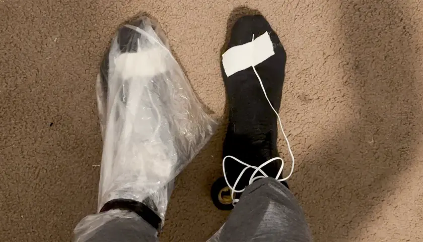 A person's feet are shown, one covered in a plastic bag, the other in a black sock with a white string and tape. These are makeshift vapor barrier liner socks for a DIY project.