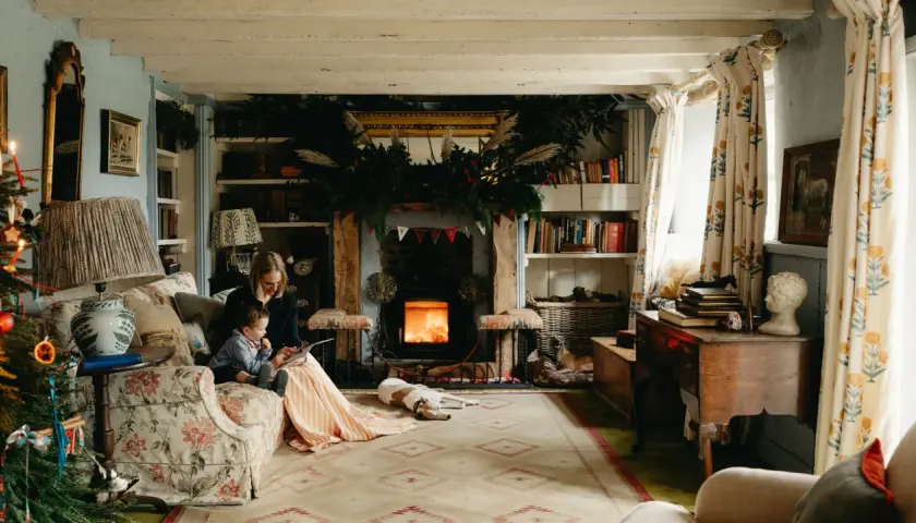 Cozy cottagecore house scene with a woman and child reading by a fireplace, dogs resting, and festive Christmas decorations.