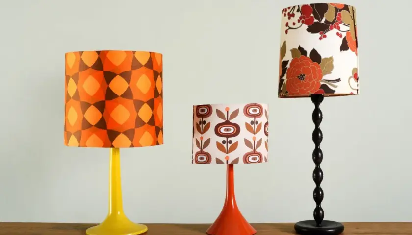 Tres lámparas de mesa con pantallas de tela de estilo retro. Una tiene un estampado geométrico naranja y marrón, otra flores marrones y naranjas sobre fondo crema, y la tercera un estampado floral en tonos rojizos y marrones.