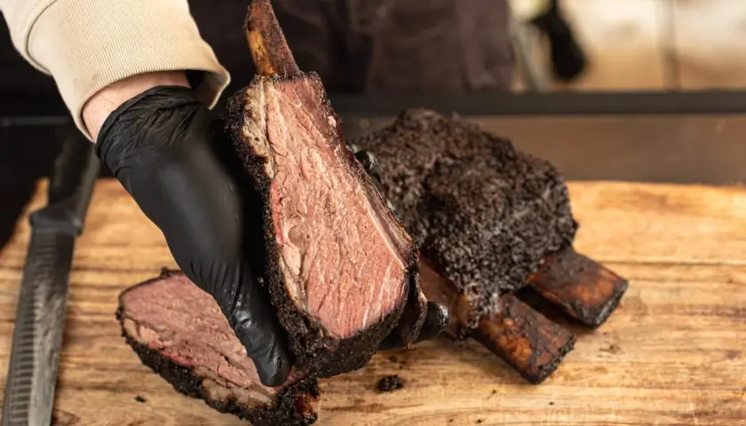 Une main gantée présente une côte de bœuf fumée, tendre et juteuse, sur une planche en bois.