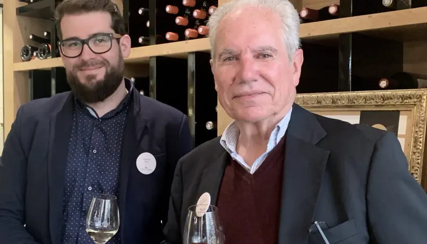 Deux hommes dégustent du vin devant des étagères remplies de bouteilles. L'un porte un badge "Domaine Rozel - Grignan-les-Adhémar avis".