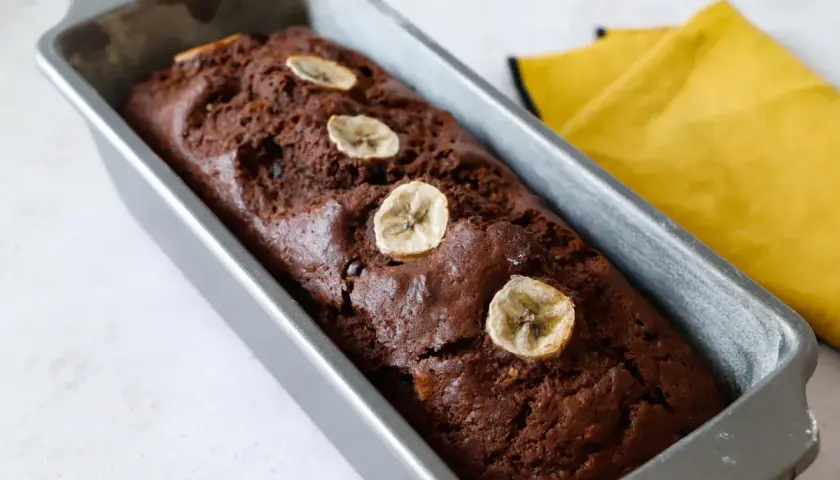 Plumcake banana e cioccolato appena sfornato, decorato con fettine di banana essiccata, in uno stampo metallico.