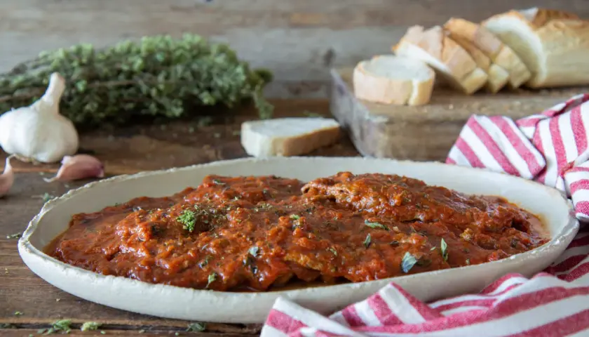 Herzhaftes Carne alla Pizzaiola in Tomatensauce, garniert mit Kräutern, neben Knoblauch, Brot und einem rot-weiß gestreiften Tuch.