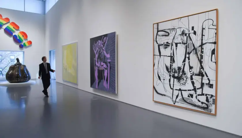 Un homme en costume observe une **analyse d'une œuvre d'art contemporain** dans une galerie. Des peintures abstraites et une sculpture colorée ornent les murs blancs.