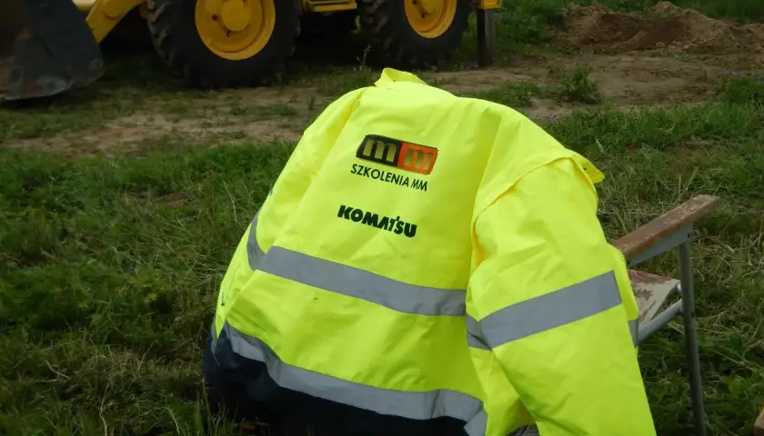 Kamizelka z logo Komatsu i napisem "Szkolenia MM" leży na krześle obok koparki. Idealna na testy na koparki jednonaczyniowe.