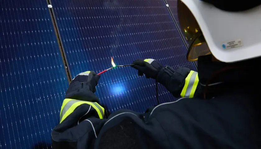 Strażak w białym hełmie i rękawicach zbliża iskrzący drut do paneli słonecznych, tworząc łuk elektryczny.