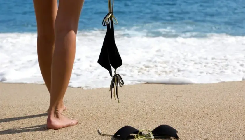 Nogi na plaży, obok leżą czarne bikini. Słońce grzeje, a fale obmywają brzeg. Czas na kąpiel dla golasy.