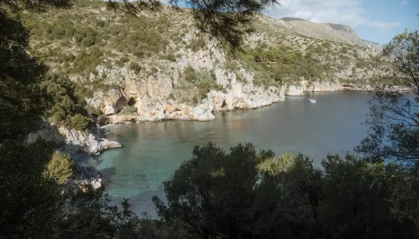 A serene view of Baia degli Infreschi, a secluded cove with clear turquoise waters, rocky cliffs, and lush greenery. A lone sailboat drifts peacefully in the bay.
