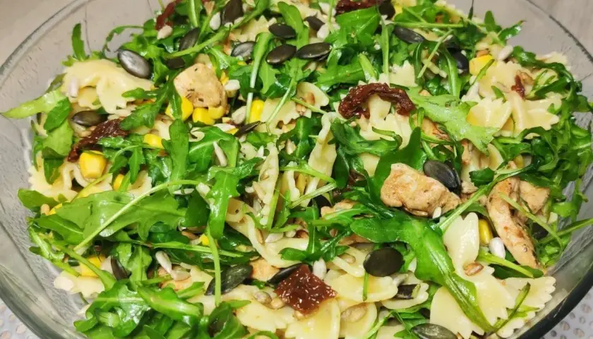 Pyszna sałatka z makaronem farfalle, rukolą, kurczakiem, kukurydzą i aromatycznymi suszonymi pomidorami. Posypana pestkami dyni i słonecznika.