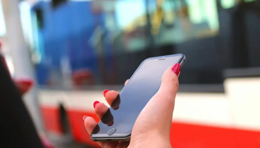 Dłoń z czerwonymi paznokciami trzyma smartfon, na którym można kupić bilet ZTM Warszawa online. W tle widać autobus.