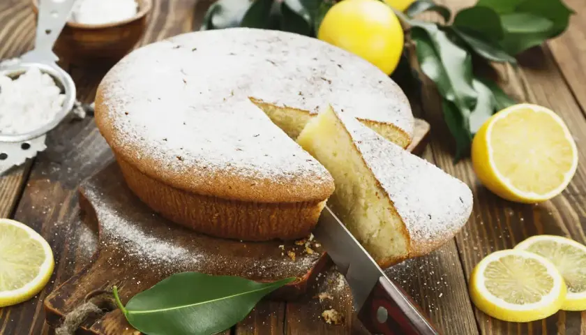 Una fetta di torta al limone morbidissima e profumatissima benedetta, cosparsa di zucchero a velo, con limoni freschi e foglie verdi su un tagliere di legno.