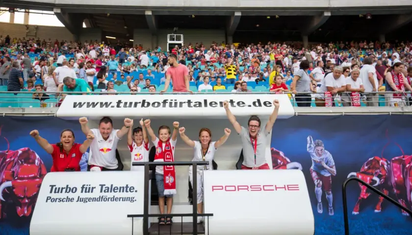Zwei Kinder und zwei Erwachsene jubeln auf einer Tribüne vor einem Banner mit Bullen und einem Läufer.