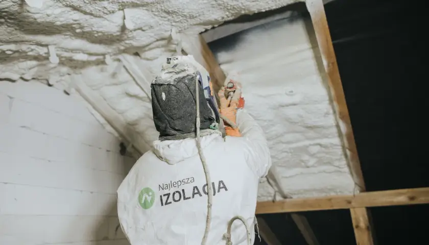 Pracownik w kombinezonie natryskuje twardą piankę poliuretanową na poddasze, tworząc skuteczną izolację.