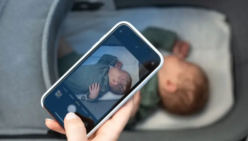 En hand håller en mobiltelefon och fotograferar en sovande bebis i en vagn. En billig babyvakt kan vara bra för att hålla koll.