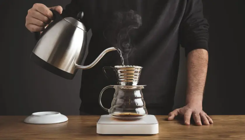 Mężczyzna przelewa gorącą wodę z czajniczka do drippera Kalita, przygotowując aromatyczną kawę.