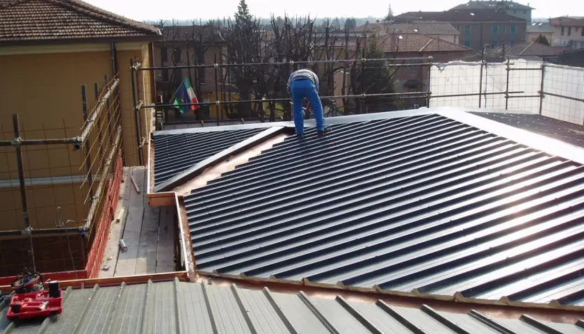 Operaio su tetto con muricci e tavelloni in fase di posa. Scaffolding e bandiera italiana sullo sfondo.