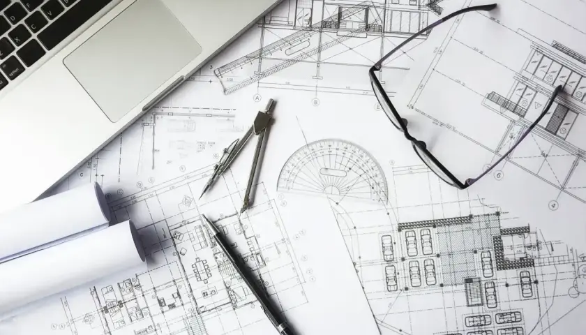 Na biurku leżą plany architektoniczne, laptop, cyrkiel, okulary i długopis. To miejsce pracy nad kosztorysem budowlanym.