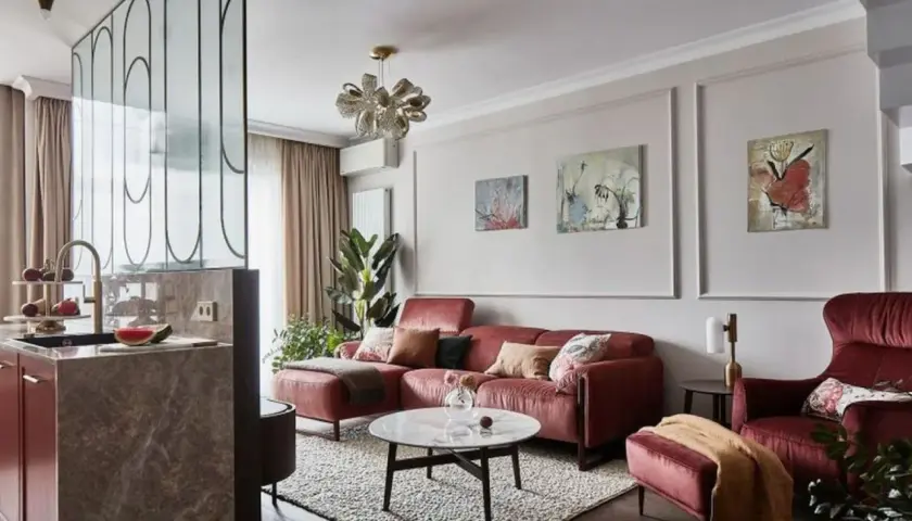 Elegancki salon z bordową kanapą i fotelem. Modne ściany w salonie zdobią obrazy, a kuchnia oddzielona jest ażurową ścianką.