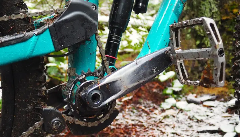 Close-up of a muddy mountain bike drivetrain with a teal frame, showing the crankset, chain, and pedal.