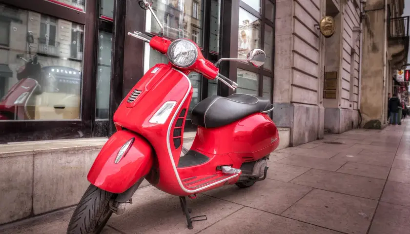 Czerwony skuter Vespa, idealny do zdobycia **prawo jazdy AM**, stoi na chodniku przed budynkiem.