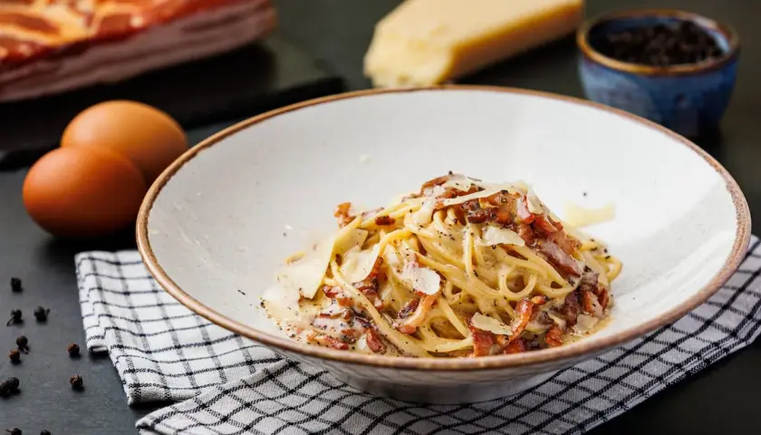 Un plato de spaghetti carbonara cremoso, con trozos de panceta crujiente y queso rallado. Ingredientes frescos como huevos, queso y pimienta negra completan la escena.