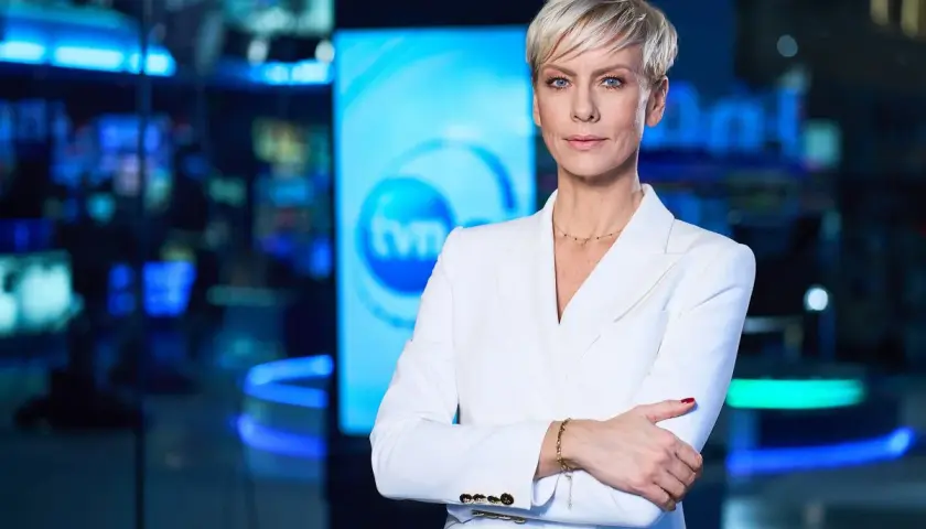 Kobieta w białej marynarce, prezenterka TVN Fakty, stoi na tle studia. Jej spojrzenie jest pewne, a postawa świadczy o profesjonalizmie.