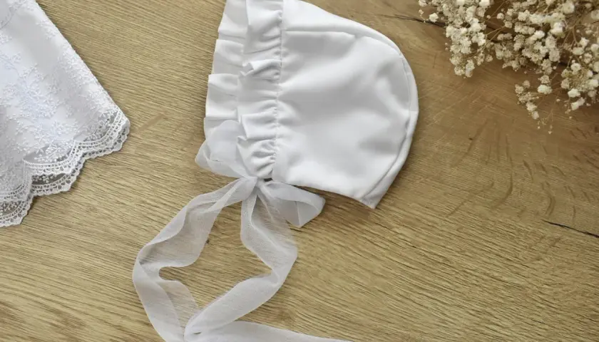 Elegancka, biała bonetka do chrztu z tiulową kokardką i koronkowym wykończeniem.