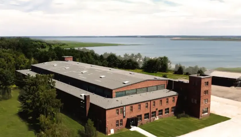 Dawny kompleks przemysłowy, dziś arche Mielno, z ceglanymi budynkami i wieżą, nad jeziorem.
